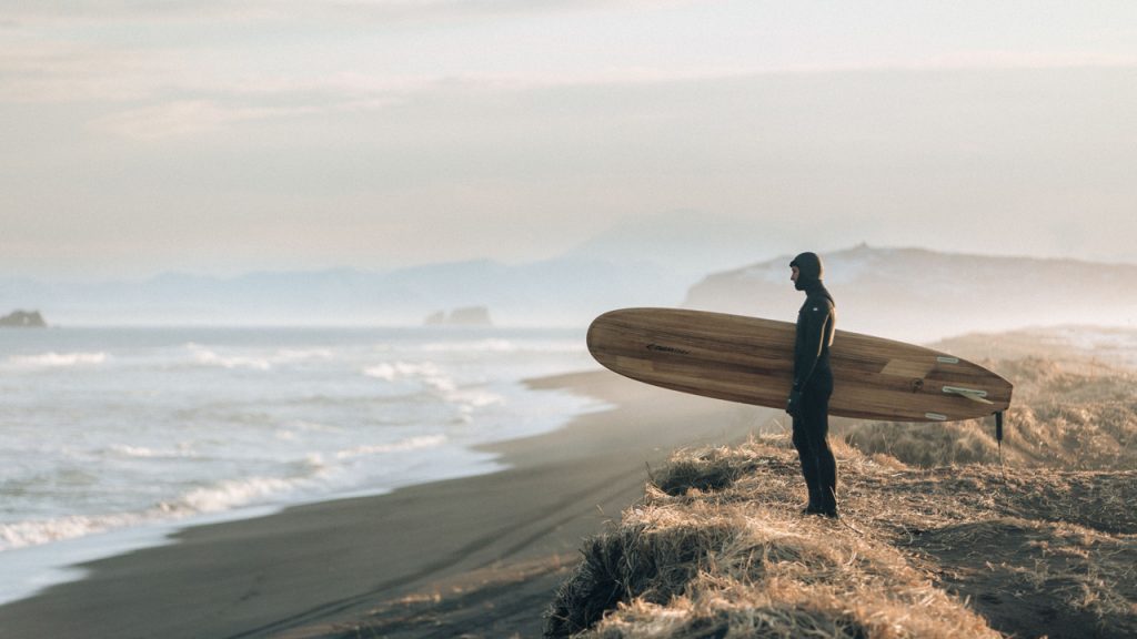 Ein Mann mit Surfbrett unterm Arm steht vor dem Meeresufer.